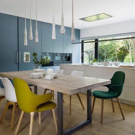 Cozinha com bancada em ilha branca, piso de madeira, armários azul, paredes em concreto aparente, mesa com cadeiras coloridas, brancas, verde claro e verde escuro.
