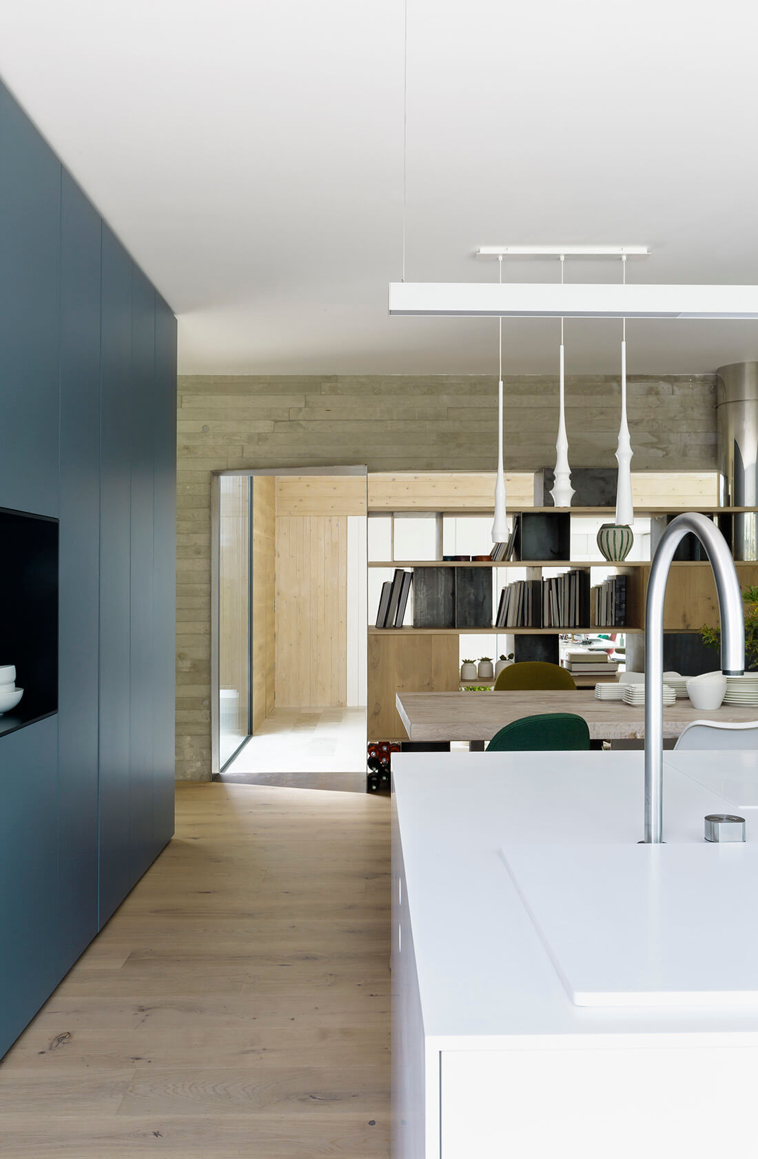 Cozinha com bancada em ilha branca, piso de madeira, armários azul, paredes em concreto aparente.