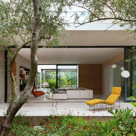 Casa Proyecto CH - Espanha. Sala de estar vista do jardim com sofa grande e cinza, poltrona couro marrom, tapete bege com preto, laje de concreto aparente, poltrona em couro marrom, chaise amarela na varanda.