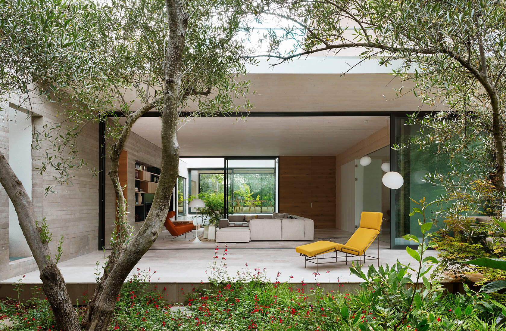 Casa Proyecto CH - Espanha. Sala de estar vista do jardim com sofa grande e cinza, poltrona couro marrom, tapete bege com preto, laje de concreto aparente, poltrona em couro marrom, chaise amarela na varanda.
