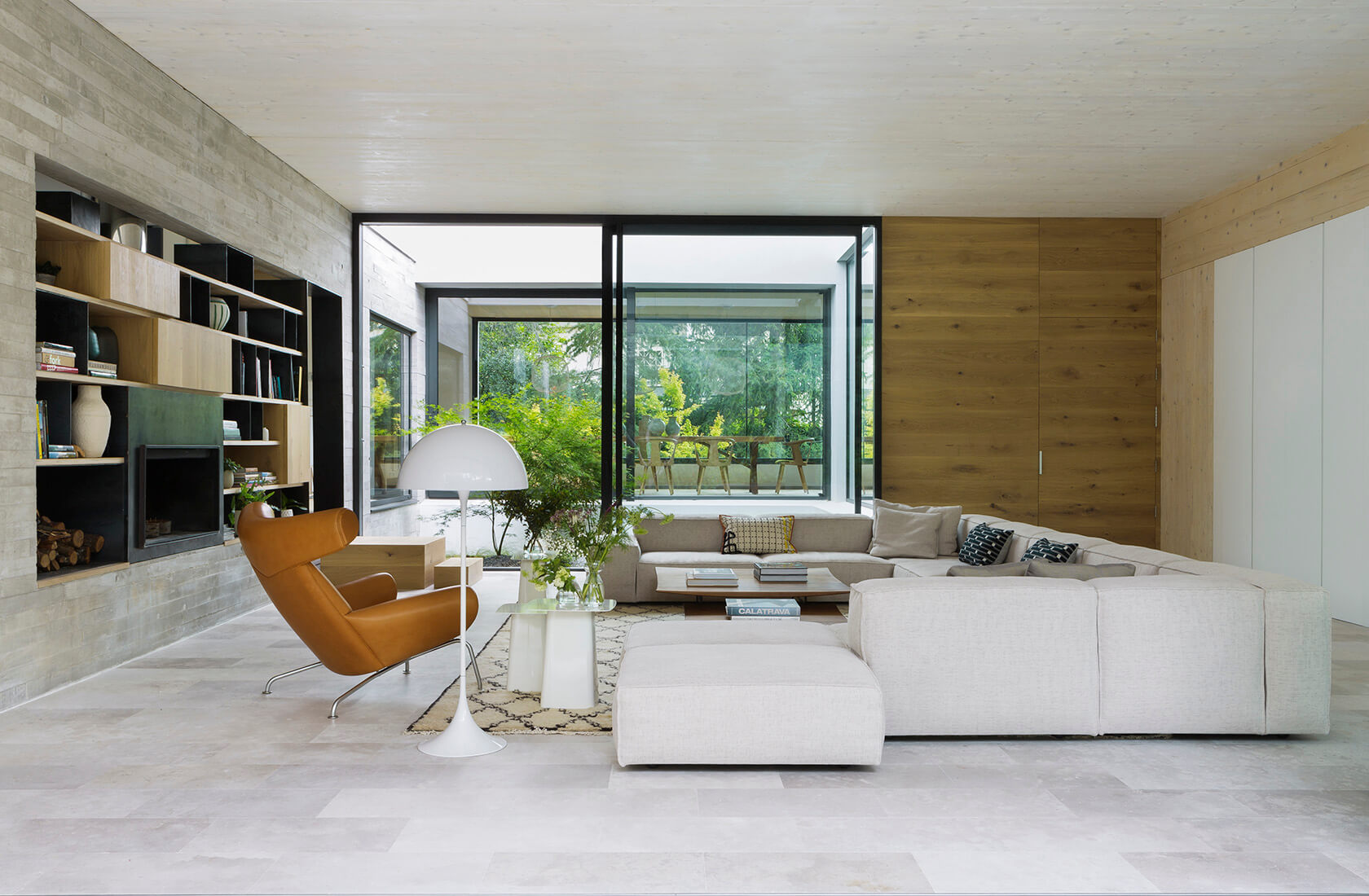 Casa Proyecto CH - Espanha. Sala de estar com sofa grande e cinza, poltrona couro marrom, tapete bege com preto, laje de concreto aparente e portas grandes do piso ao teto com vista para a natureza.