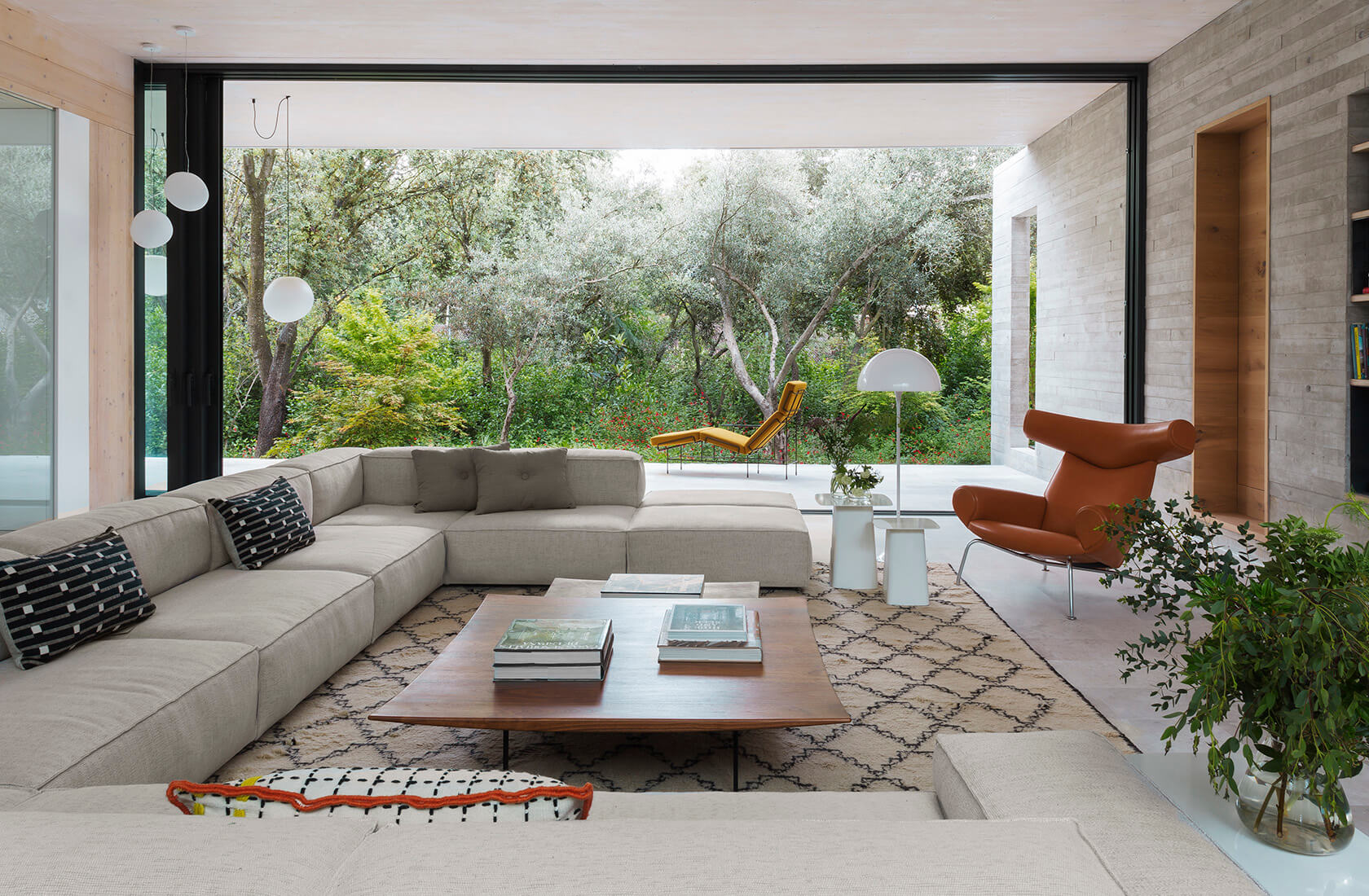 Casa Proyecto CH - Espanha. Sala de estar com sofa grande e cinza, tapete bege com preto, laje de concreto aparente e portas grandes do piso ao teto com vista para a natureza.